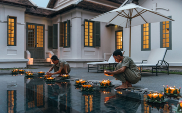 Two guests seated at the edge of a plunge pool at Amantaka, with the colonial-style building reflected in the water.
