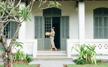 A guest stands on the front veranda of a Mekong Pool Suite at Amantaka, framed by white columns and tropical plants.