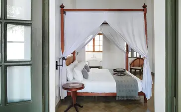 Canopied bed with white linens in a principal bedroom at Amantaka, Laos, framed by open wooden doors.