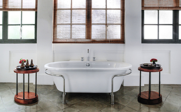 Freestanding bathtub with vintage pedestal sink at Amantaka, flanked by decorative side tables beneath tall windows.