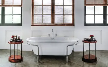 Freestanding bathtub with vintage pedestal sink at Amantaka, flanked by decorative side tables beneath tall windows.