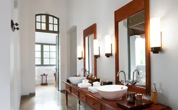 Amantaka suite bathroom with wooden-framed mirror, dual vanity and natural light from window.