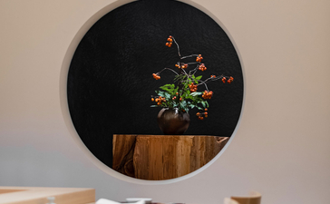 A circular black mirror with wooden frame reflects green foliage, mounted above a wooden dining table at Aman Tokyo.