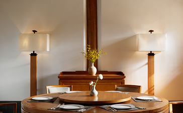 Wooden dining table beneath a brass pendant light at Amandayan, with exposed beam ceiling and upholstered chairs.