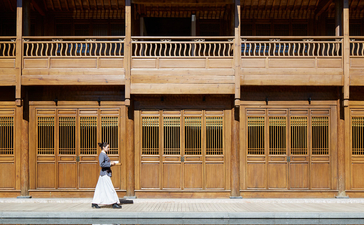 Woman in white dress seated before traditional wooden architecture at Amandayan, reflected in still water.