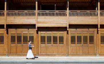 Woman in white dress seated before traditional wooden architecture at Amandayan, reflected in still water.