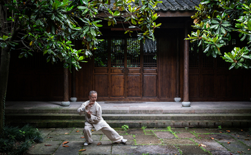 Tai chi practitioner in white clothing performing a stance in the courtyard at Amanfayun, with traditional wooden doors and green vines framing the scene.