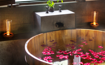 Wooden soaking tub filled with rose petals and candles at Amanfayun, with potted plant and window beyond.