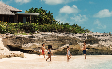 Family wading in turquoise waters at Amanyara's beachfront with rocky outcrops and accommodation visible above.