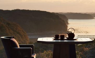A dining table set for two overlooks a serene coastal landscape at sunset, Amanemu resort.