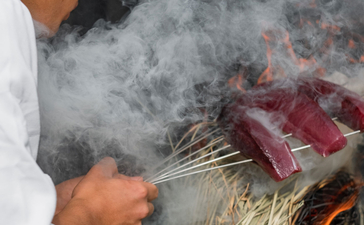Chef grilling meat over flames at Amanemu resort, smoke rising from the grill.
