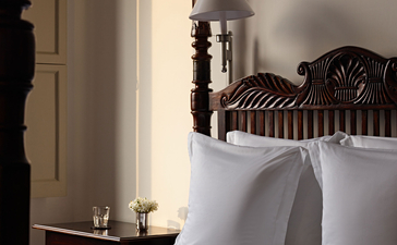 Ornate wooden bed with white linens and pillows at Amangalla, with candlelit bedside table and arched doorway.