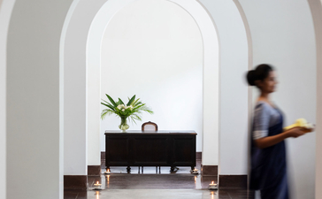 Arched corridor at Amangalla with polished grey stone floor, white walls, and a figure walking past a potted plant on a dark console table.