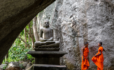 Stone steps descend beneath a cave overhang at Amangalla, with two monks in orange robes standing nearby.