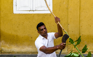 A man in white clothing sits against a golden-yellow wall at Amangalla, holding a long wooden staff upright.