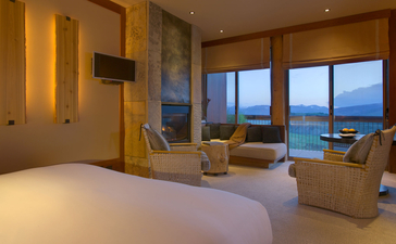 Amangani Superior Suite with floor-to-ceiling windows overlooking mountain views, featuring a principal bedroom and seating area bathed in warm evening light.