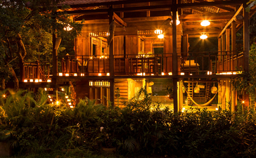 Amansara pavilion illuminated at dusk, warm golden light glowing through wooden shutters onto manicured gardens.