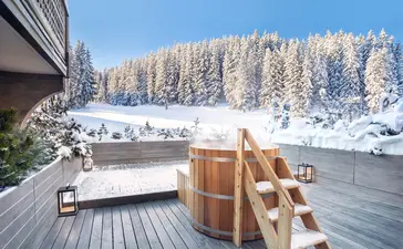 Wooden hot tub on a deck at Aman Le Mélézin overlooking snow-covered alpine forest and mountains.