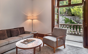 Sitting area in Milocer Garden View Suite at Aman Sveti Stefan, with grey sofa, lamp, and window overlooking gardens.