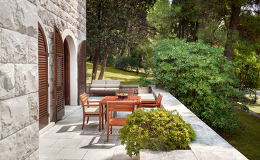 Wooden dining furniture on a stone terrace at Aman Sveti Stefan, overlooking lush greenery.