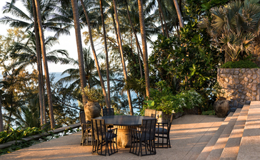 Seating area with wooden chairs on a terrace at Amanpuri resort, Thailand, framed by tall palm trees and ocean views.
