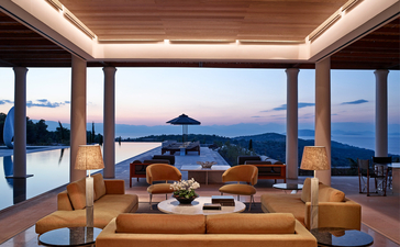 Villa lounge at Amanzoe resort at dusk, overlooking the Aegean Sea through floor-to-ceiling windows.