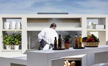 Outdoor bar area at Amanzoe resort with staff member preparing drinks, white architectural design and potted plants.
