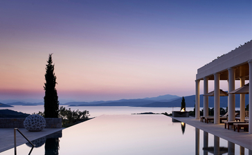 Waterfront view at Amanzoe resort at dusk, with a lighthouse and columned structure reflected in calm waters.
