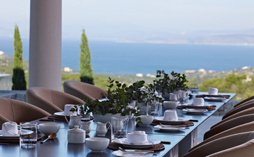 Dining terrace at Amanzoe resort overlooking the Aegean Sea, with cypress trees and coastal views.