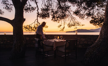 Silhouetted figures seated at a dining table beneath a pine tree at Aman Sveti Stefan, overlooking the sea at dusk.