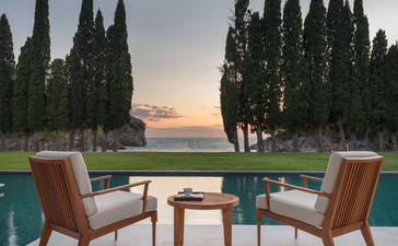 Two mid-century chairs beside a wooden table on a terrace overlooking a pool and cypress trees at sunset, Aman Sveti Stefan.