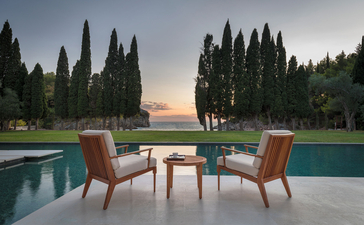Two chairs and table beside swimming pool at Aman Sveti Stefan at sunset, with cypress trees beyond.