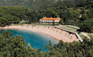 Aerial view of Aman Sveti Stefan's sandy beach cove with turquoise waters and orange-roofed buildings nestled among green hillside.