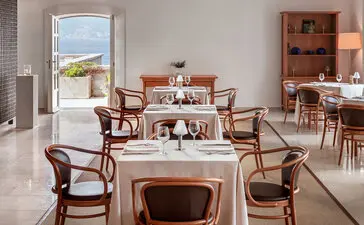 Dining area at Aman Sveti Stefan with wooden chairs, tables, and coastal views through open doors.
