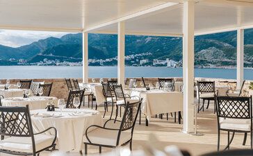Dining room at Aman Sveti Stefan with floor-to-ceiling windows overlooking the Adriatic Sea and coastal mountains.