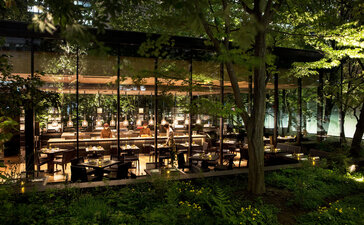 Aman Tokyo's dining café illuminated at dusk, with warm lights glowing beneath verdant trees.