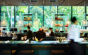 Aman Tokyo's dining café with floor-to-ceiling windows overlooking greenery, featuring a sleek counter and suspended pendant lights.