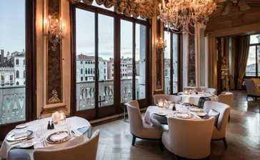 Elegant dining room at Aman Venice with canal views through tall windows, round tables set for service, and chandelier lighting.