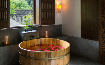 Wooden soaking tub filled with flower petals in a spa treatment room at Amanfayun, with natural light from traditional wooden shutters.