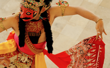 Woman in red batik dress reclining on cream bedding at Amanjiwo, surrounded by traditional Indonesian textiles and cushions.