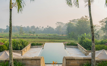 Two wooden chairs facing a plunge pool at Amanjiwo, flanked by palm trees overlooking distant temples.