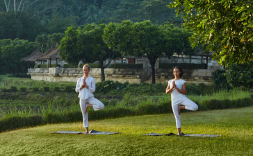 Two people practising yoga on a grass lawn at Amanjiwo, with lush greenery and a building visible across the water beyond.