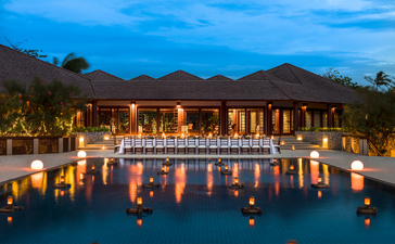 Amanpulo's dining venue illuminated at dusk, reflected in still water with mountain backdrop.