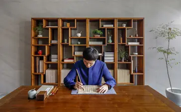 Woman in blue shirt seated at wooden desk in front of built-in shelving at Amanyangyun.