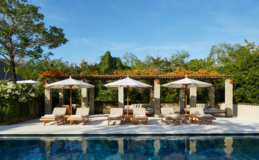 Poolside cabanas with cream canvas awnings at Aman Villas at Nusa Dua, reflected in still water beneath clear sky.