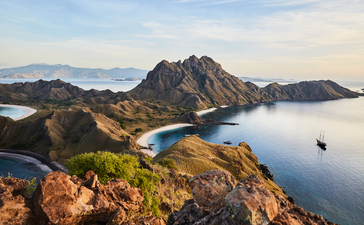 Aerial view of Amandira's rugged coastline with forested peaks meeting calm blue waters and distant mountains.