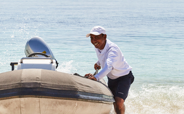 A man in a cap pulls a dinghy ashore at Amandira, Indonesia, with a sailing vessel anchored offshore.