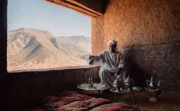 Homme assis dans une chambre d'Amanjena, contemplant les montagnes désertiques par une fenêtre.