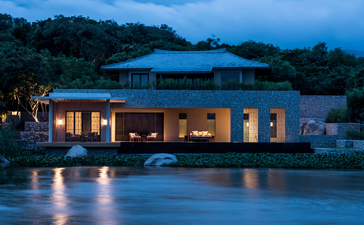Amanoi villa with illuminated interior reflected in still water at dusk, surrounded by forested landscape.