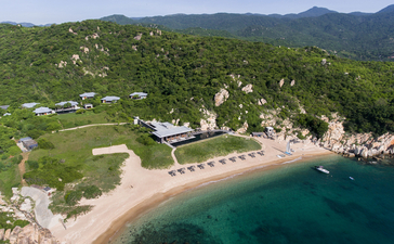 Aerial view of Amanoi's clifftop resort overlooking a sandy beach and turquoise waters, surrounded by lush hillside vegetation.